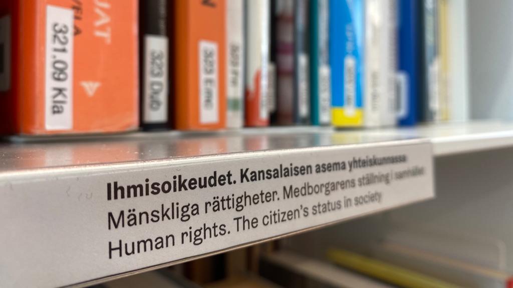 Kuvassa on kirjaston kirjahylly. Edessä kyltti, jossa luee "Ihmisoikeudet. Kansalaisen asema yhteiskunnassa.". Taustalla kirjoja.