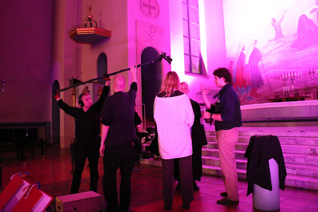 Elokuvan ohjaaja keskustelee näyttelijöiden kanssa kirkon alttarilla ääniosaston testatessa puomin paikkaa.