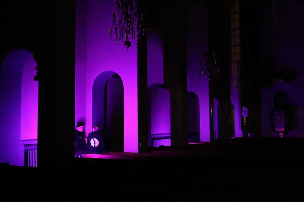 Kaksi ihmistä seisovat pimeässä kirkon käytävässä violetin valon loisteessa.