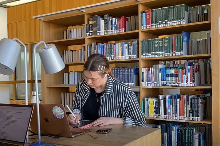 Nainen kirjoittaa kynällä kirjahyllyn edessä tietokoneen takana