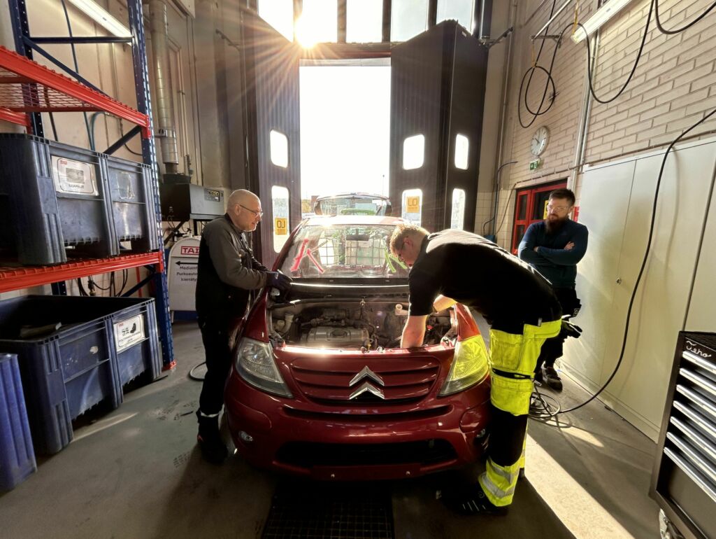 Kuvassa kolme työasuihin pukeutunutta miestä työskentelee punaisen auton äärellä hallissa. Takana on avoinna oleva hallin ovi, josta tulee sisään valoa.