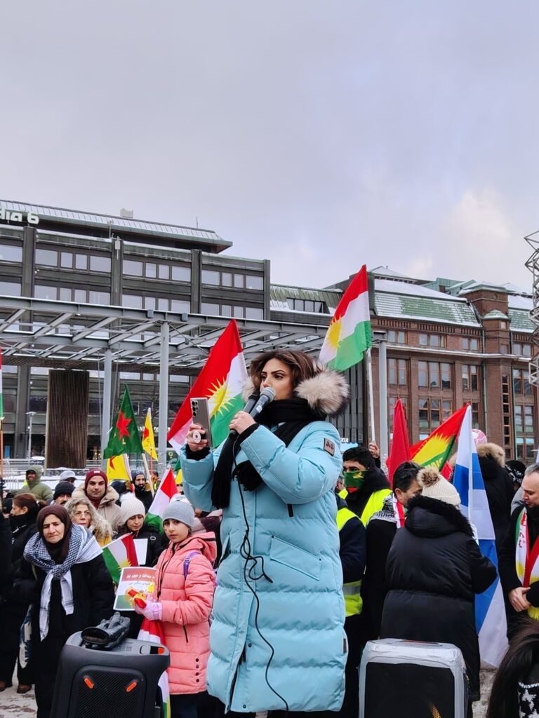 Tawar Salari puhumassa mielenosoituksessa Helsingissä. Taustalla Iranin lippuja sekä muita mielenosoittajia. 