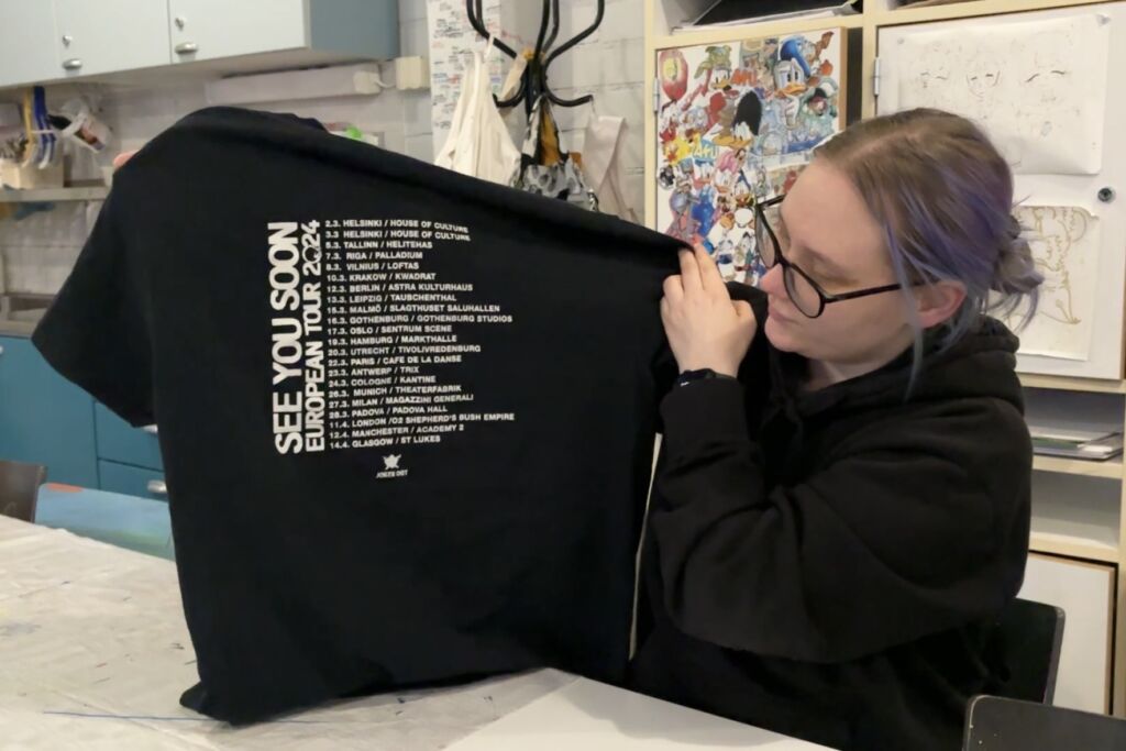 Haastateltava pitää käsissään fanipaitaa, jossa on kiertueen päivämääriä.