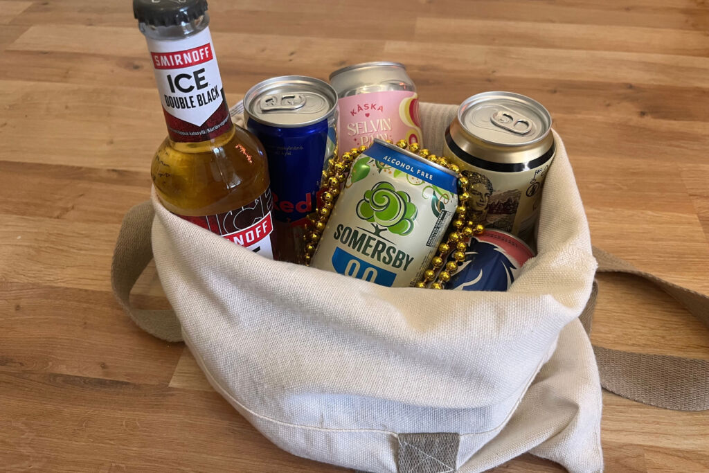 Kuvassa on kangaskassi, jonka sisällä on 6 juomaa. Yksi lasipullo ja viisi tölkkiä. Juomat ovat alkoholillisia ja alkoholittomia. Juomia ympäröi kultainen helmikoru.